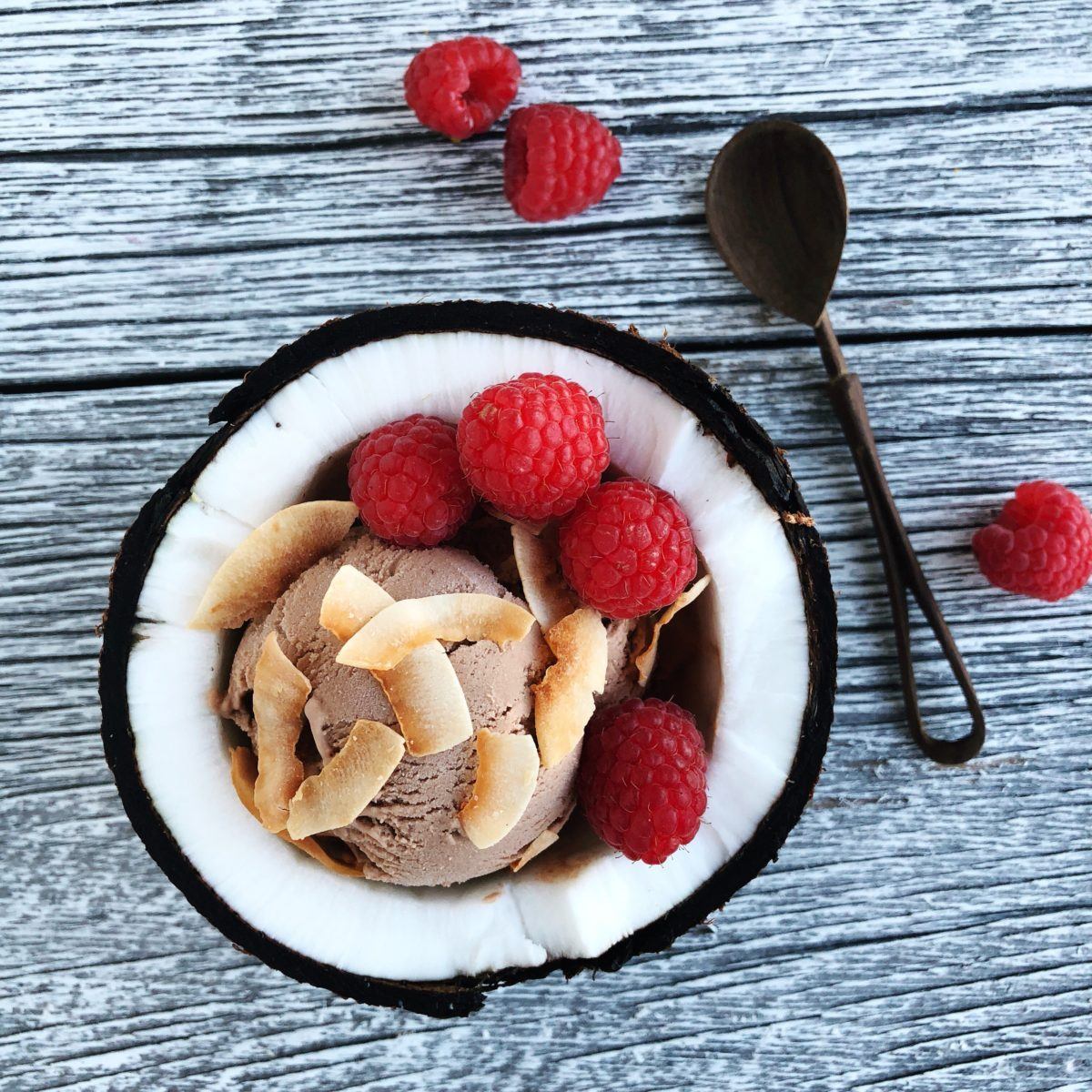 Vegan chocolate ice cream topped with toasted coconut shavings and raspberries in a half-coconut bowl, next to a wooden spoon on a painted white wooden tabletop.