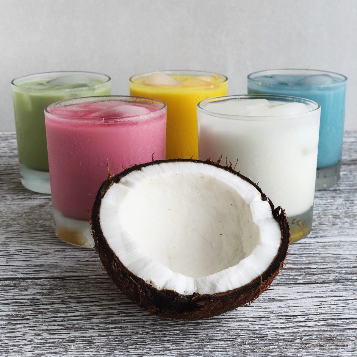 Half a coconut in front of five glasses of coconut milk, colored with beet root powder, matcha, turmeric, and spirulina