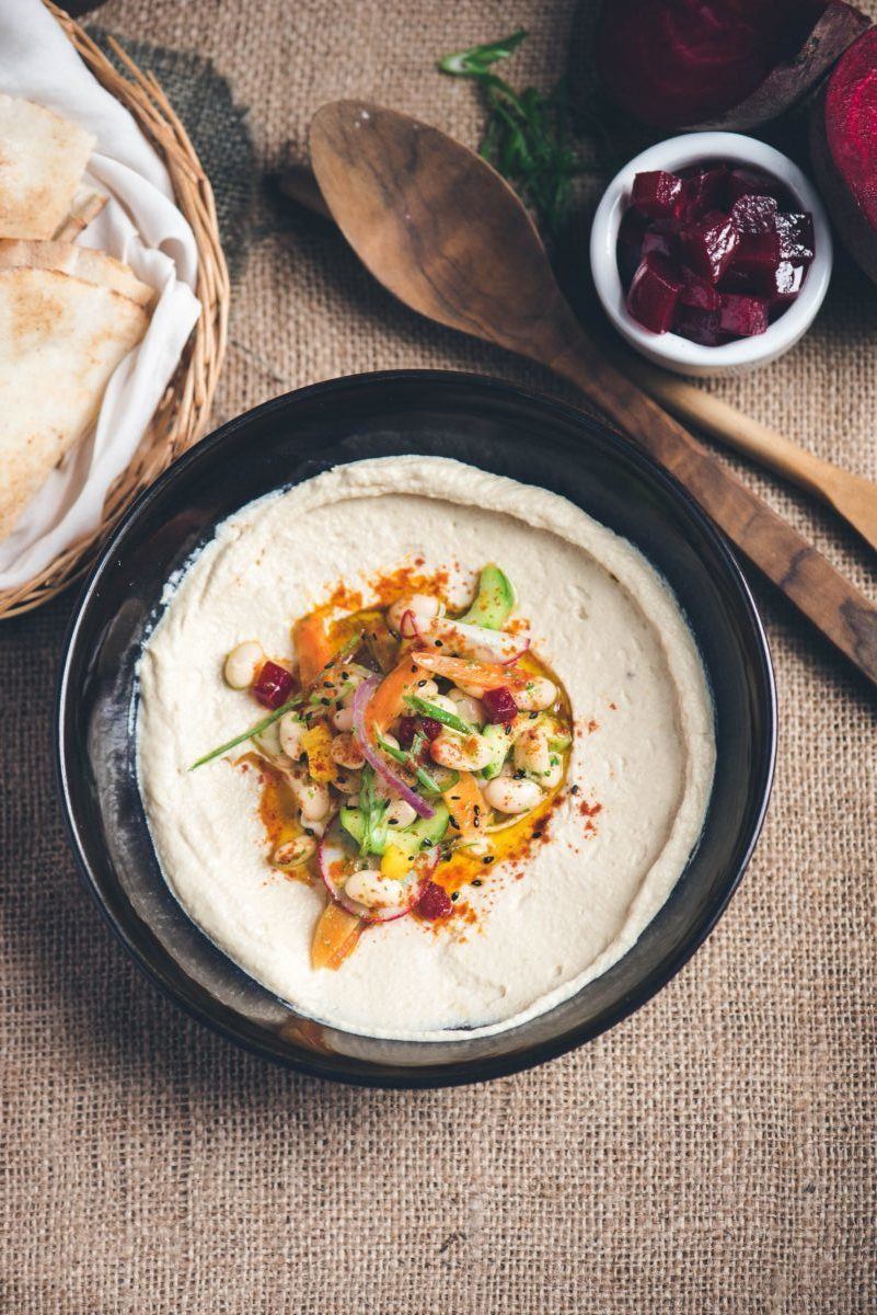 Homemade hummus made with tahini in a black bowl topped with white beans, chopped vegetables, and paprika, on a table setting with flat bread, wooden spoons, and chopped beets.