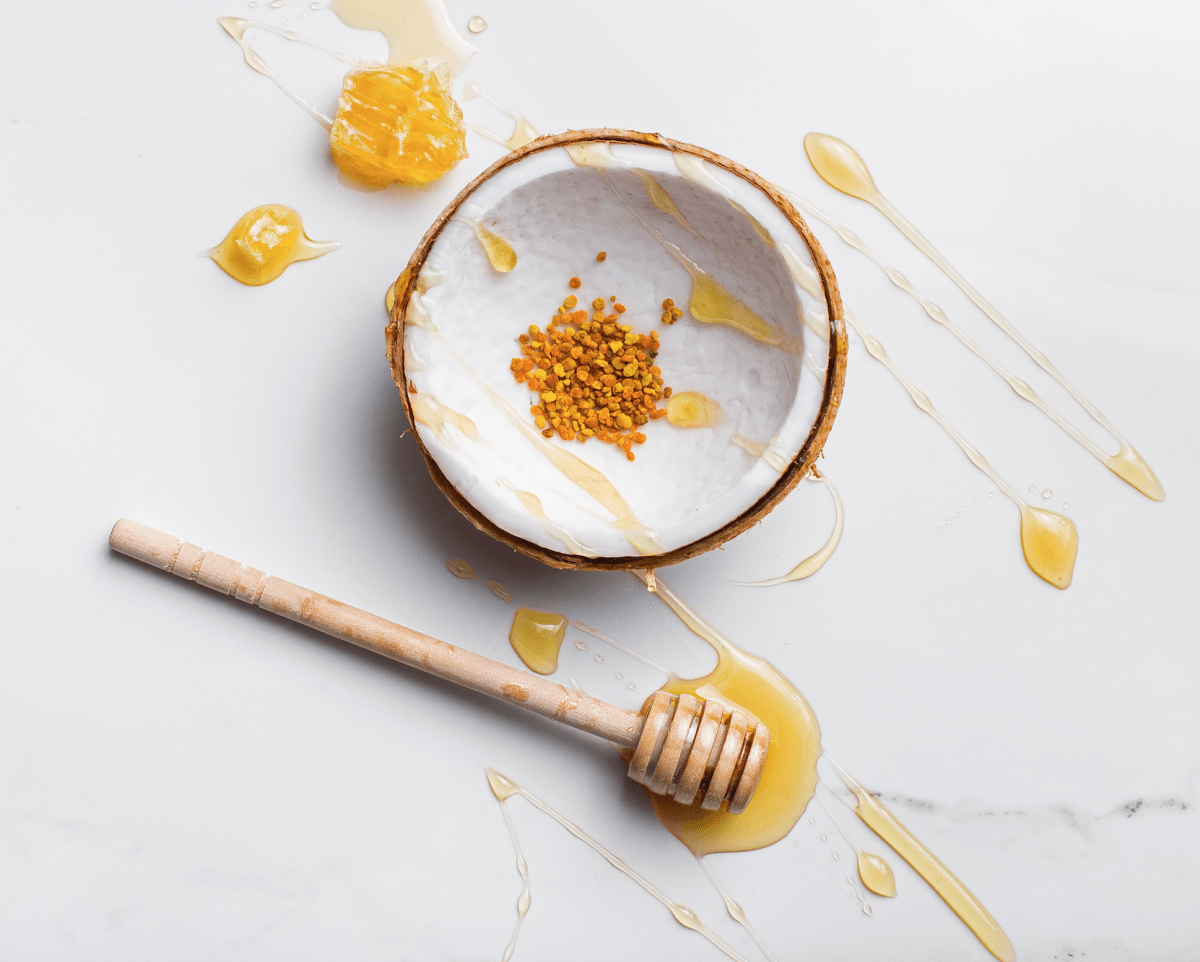A scoop of bee pollen in half a coconut on a white background, surrounded by raw honeycomb, raw honey drizzle, and a wooden honey dripper. 