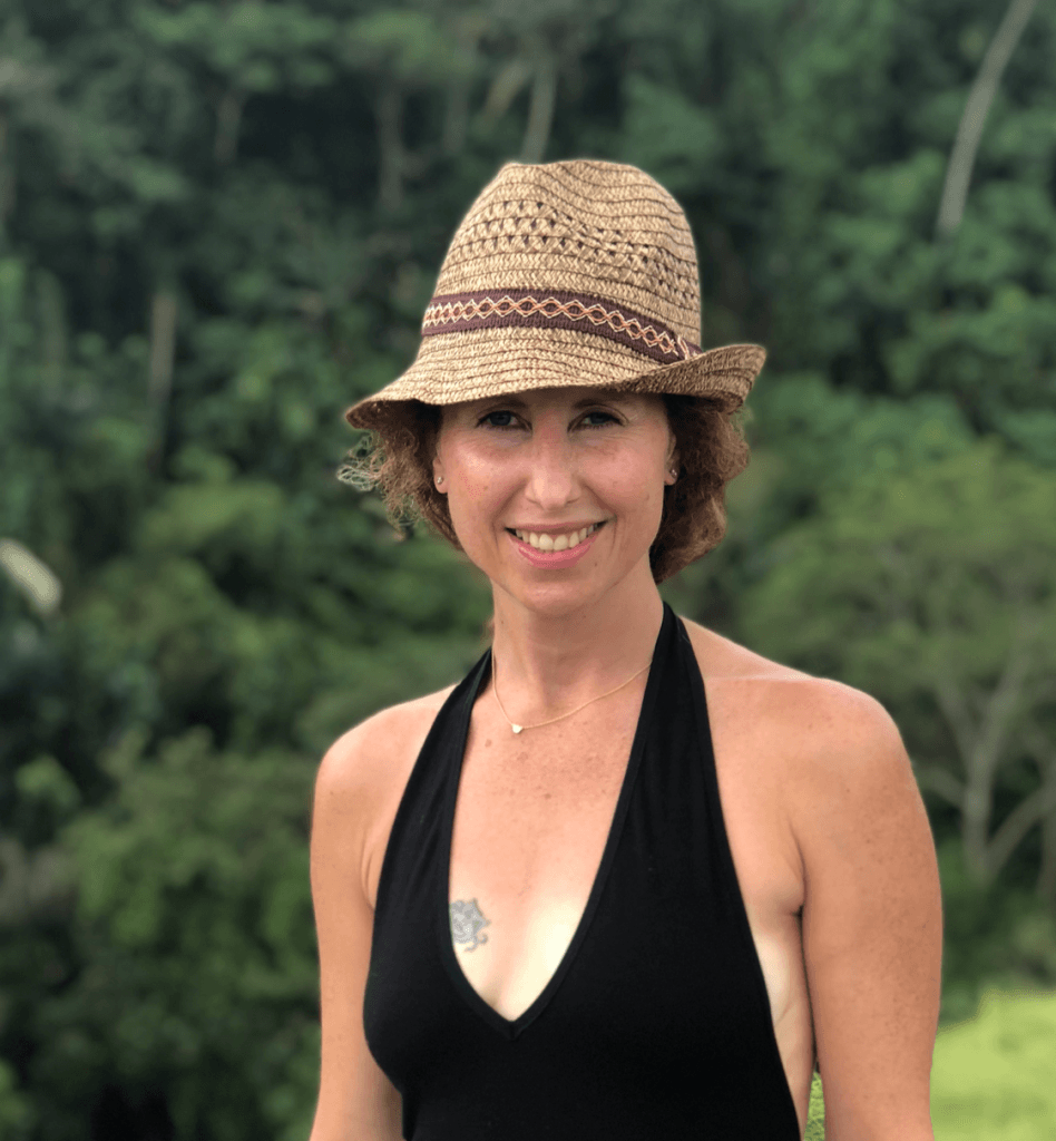 A woman smiles standing in front of a tropical rainforest wearing a sunhat. 