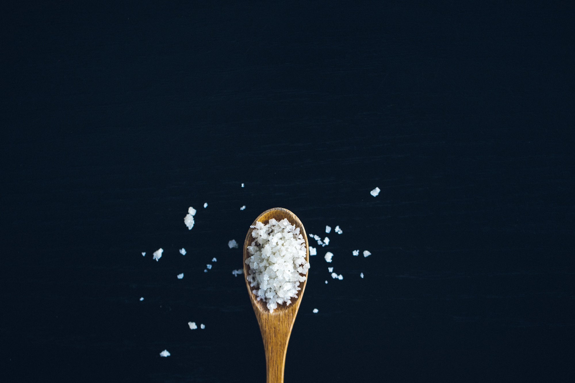 A wood spoon of crisp white sea salt on a dark blue background with sprinkles of salt. 