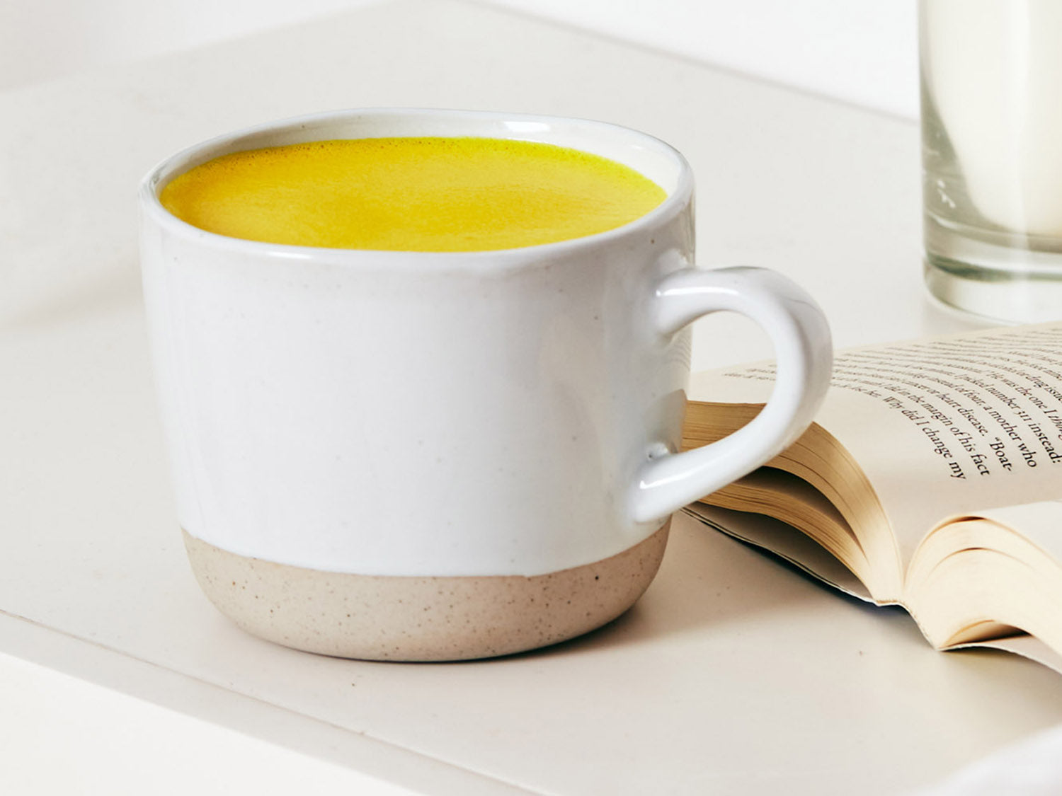 White mug of Restore turmeric latte sitting on a bedside table with an open book and candle beside it