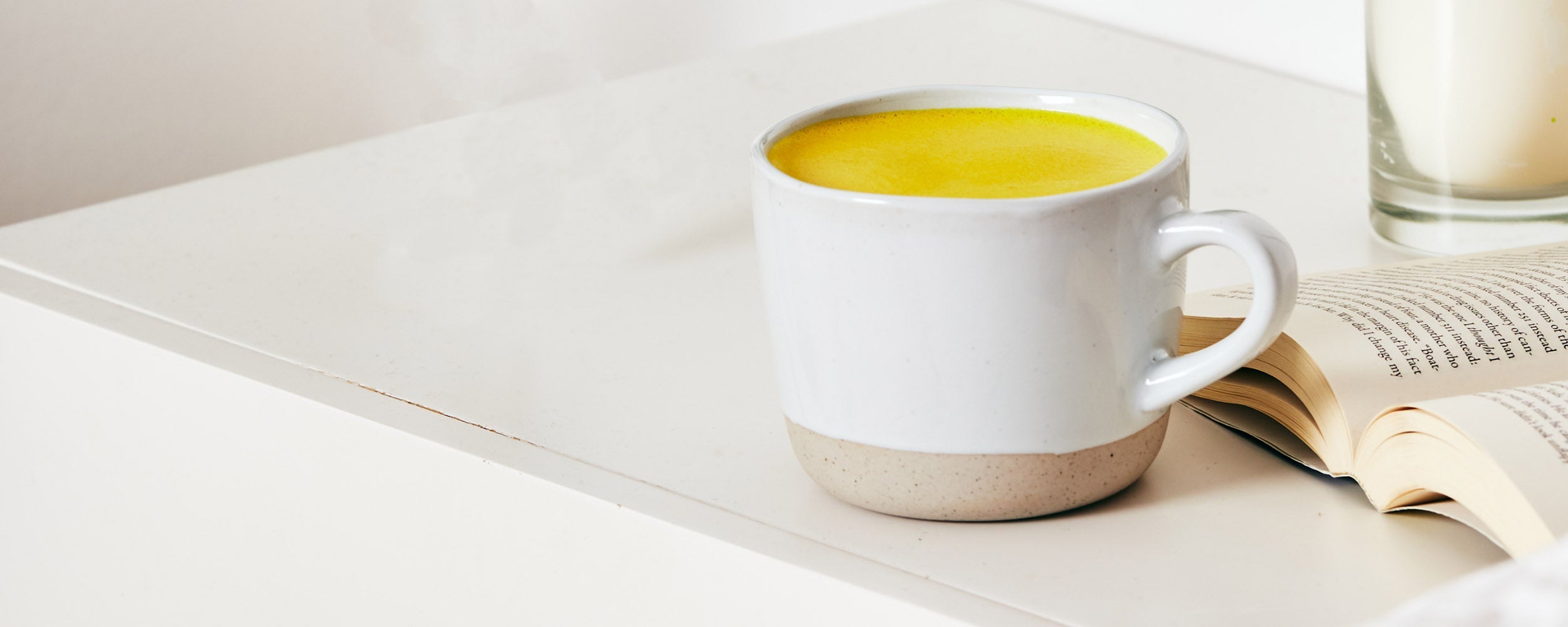 White mug of Restore turmeric latte sitting on a bedside table with an open book and candle beside it