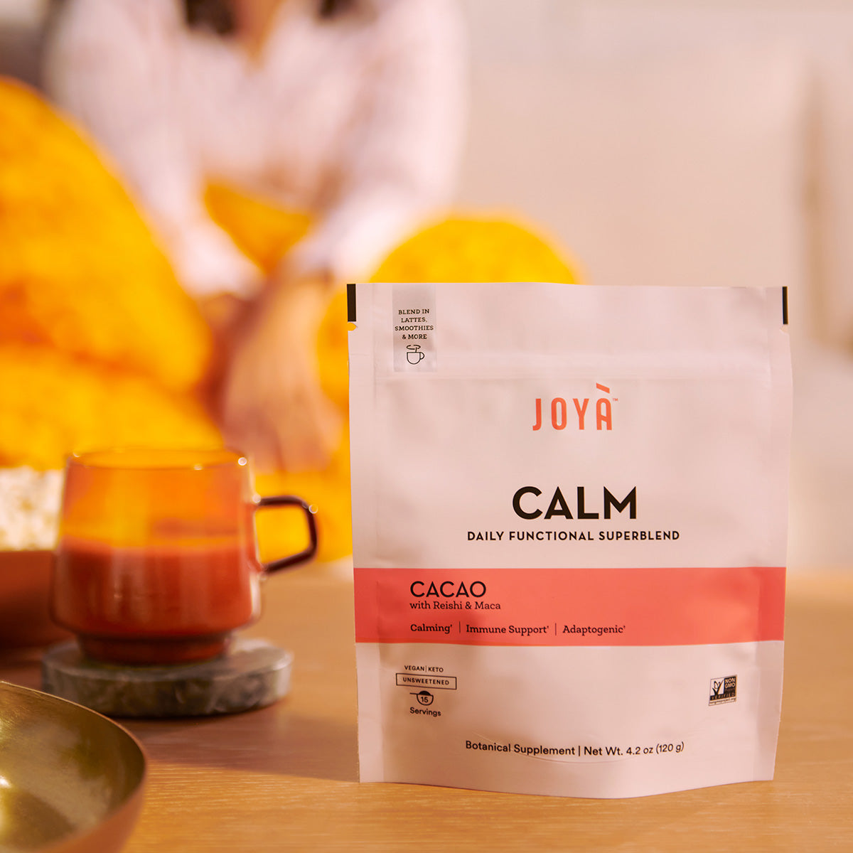 Calm Superblend 15-serving pouch beside a mug of hot Calm cacao latte while a young woman relaxes on the couch in the background