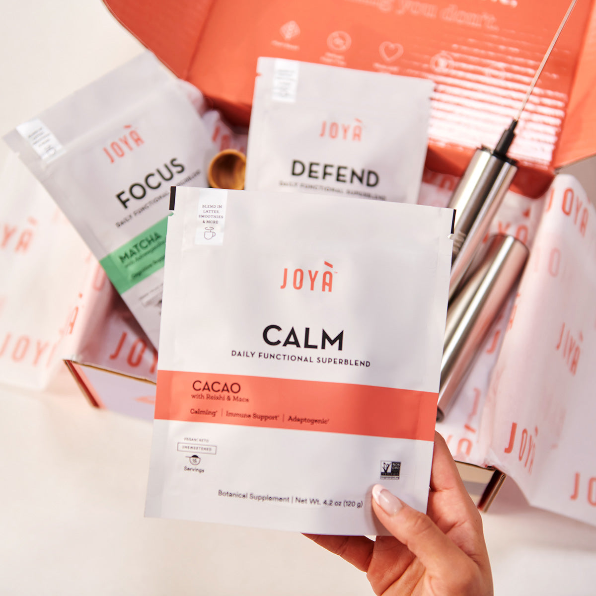 Young woman's hand holding a Calm Superblend 15-serving pouch above the JOYÀ Subscription Box Starter Bundle with Focus Superblend, Defend Superblend, wooden spoon and rechargeable frother in the box on a white tabletop
