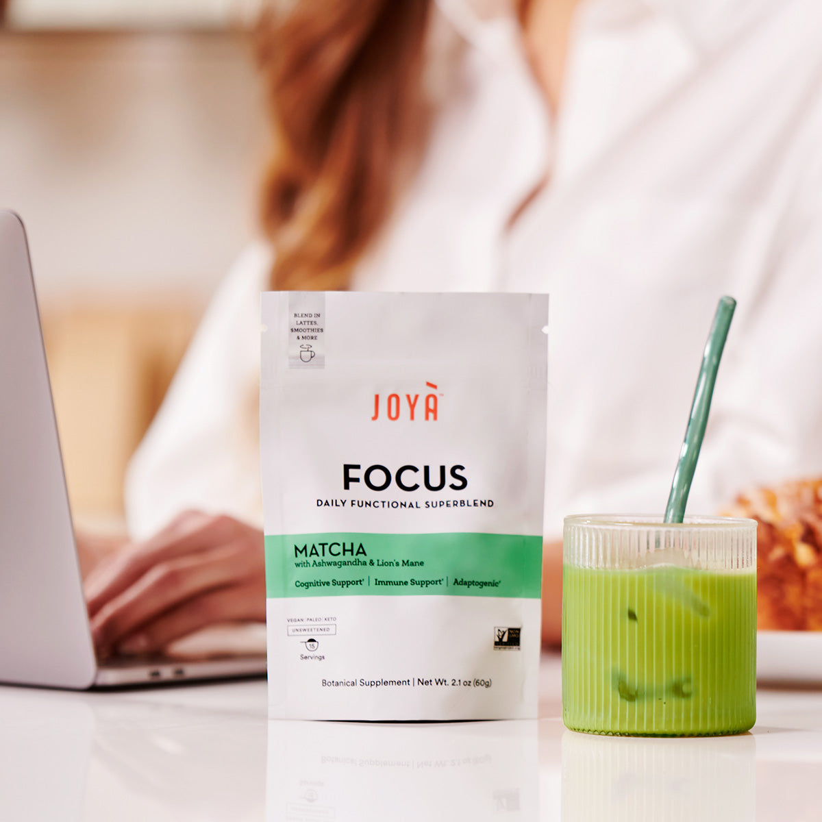 Focus Superblend 15-serving pouch beside a cup of iced Focus matcha latte while a young woman works on her laptop in the background
