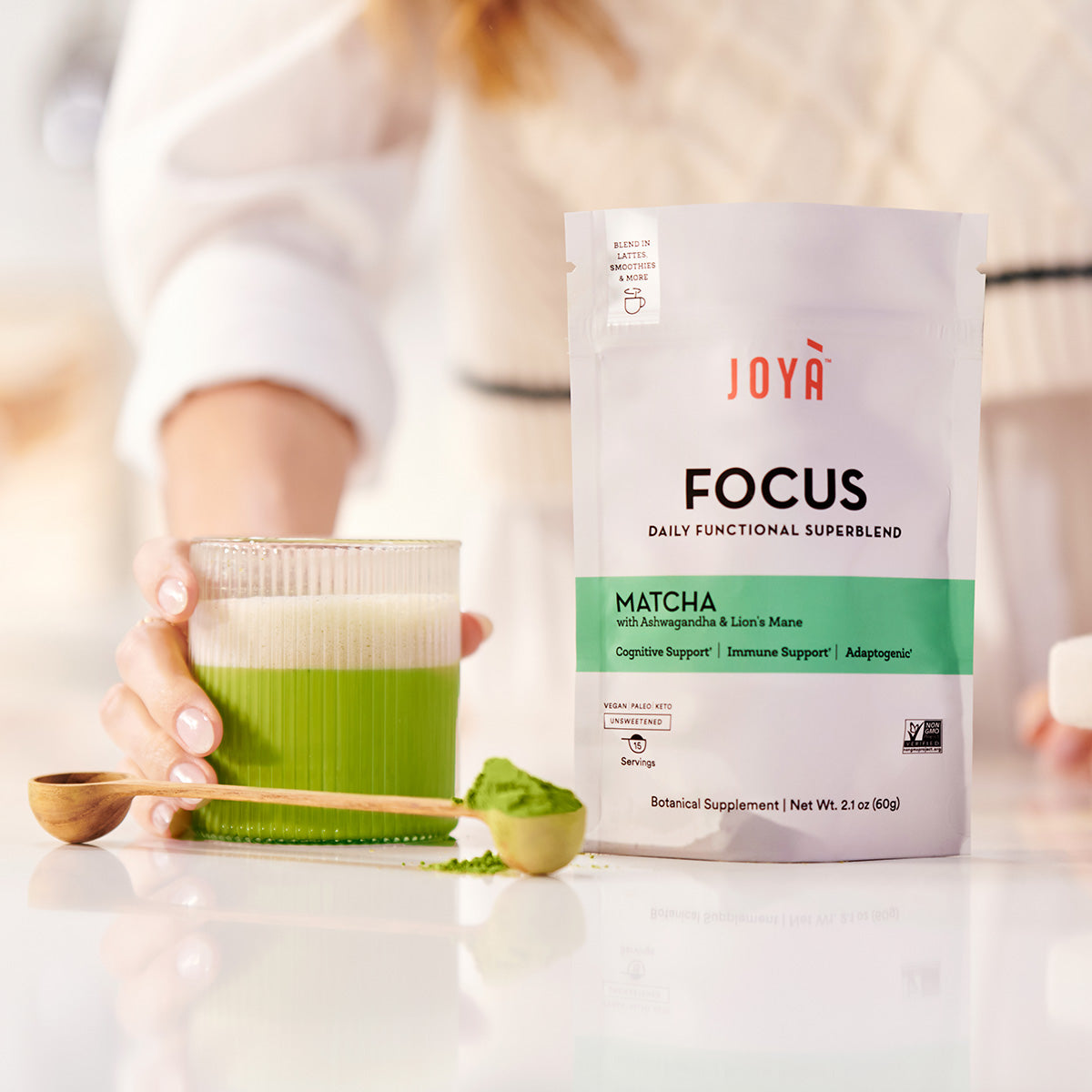 Focus Superblend 15-serving pouch beside a wooden spoon and a clear glass of Focus matcha latte being held by a young woman standing in her kitchen