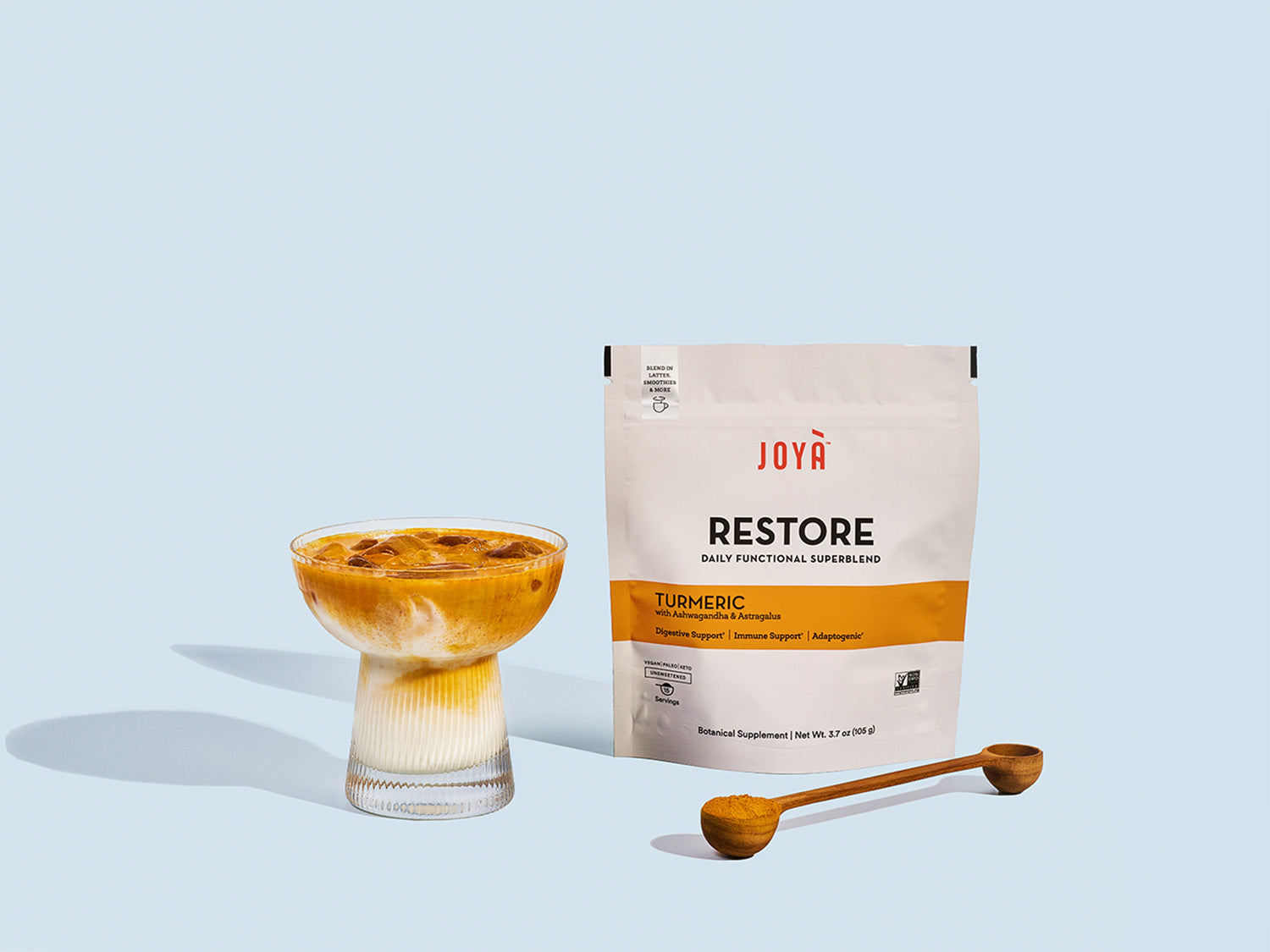 Restore Superblend 15-serving pouch sitting beside a wooden spoon and iced Restore turmeric latte in a clear glass on a blue background