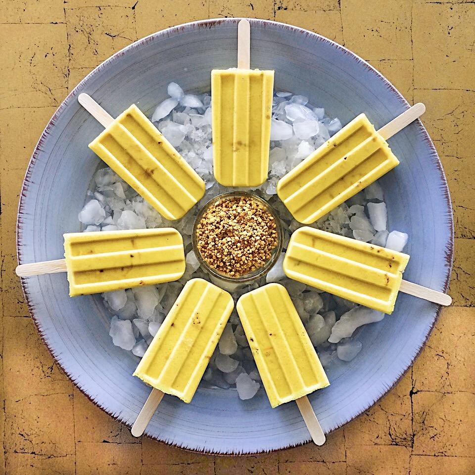 Dairy-free golden milk popsicles rest on a bed of ice on a blue plate in a circle, with a glass bowl of bee pollen in the center. 