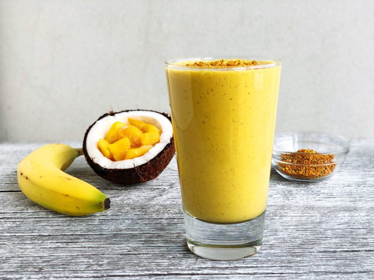 A tropical turmeric smoothie in a tall glass topped with bee pollen, next to a banana, coconut half filled with chopped mango, and a small bowl of bee pollen.