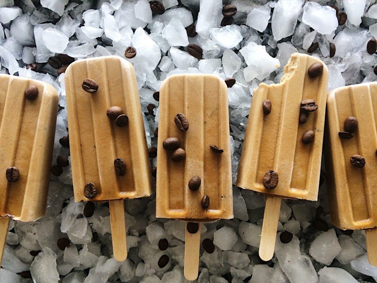 Vegan and dairy-free cold brew tahini popsicles lie on a bed of ice with coffee beans sprinkled on top.
