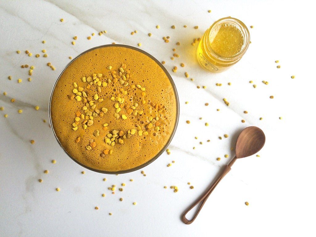 Aerial view of a tropical smoothie topped with bee pollen, surrounded by sprinkled bee pollen, a wooden spoon, and a glass jar of raw honey. 