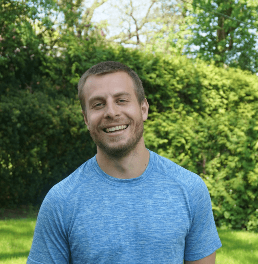 Young fit man in a blue t-shirt stands smiling outside in front of a green hedge. 