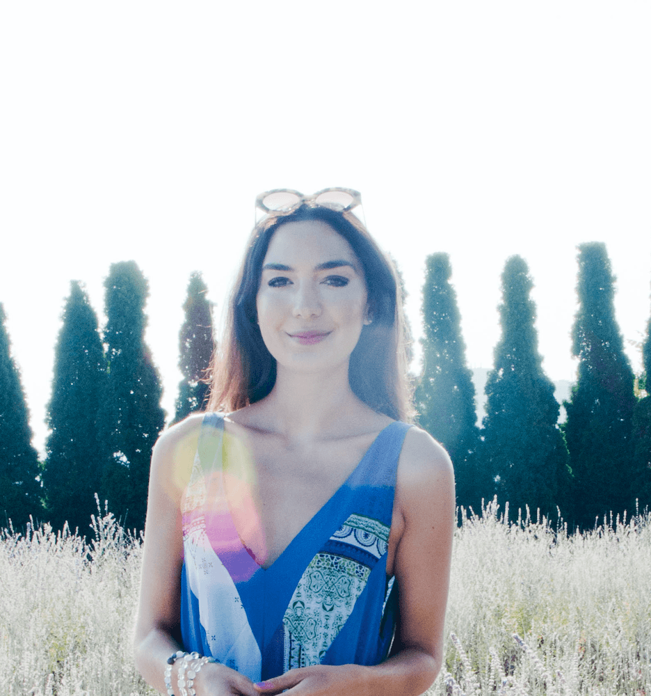 Young woman, Kiki Athanassoulias, stands outside in a sunny field in front of a row of trees. 
