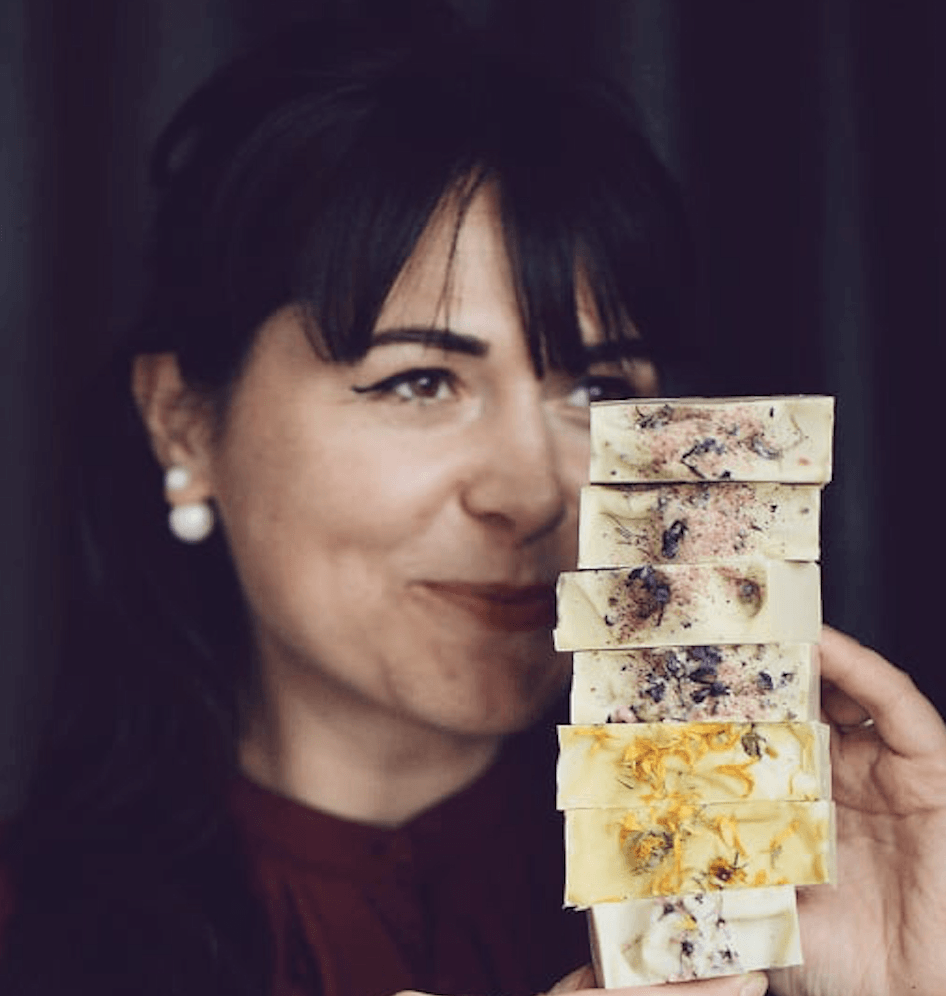 A woman, Natalia Vanthienen, with dark brown hair against a dark background holding a stack of homemade natural soaps. 