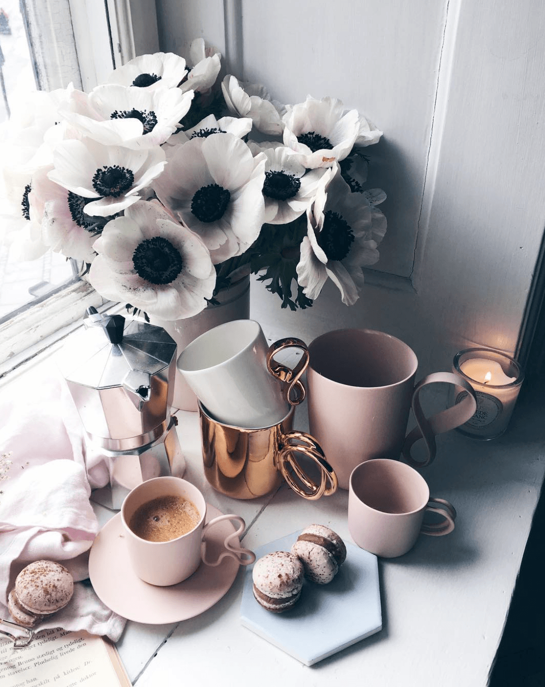 Pink coffee cups on pink saucers sit on a windowsill surrounded by vanilla macarons and next to a vase full of big white flowers.