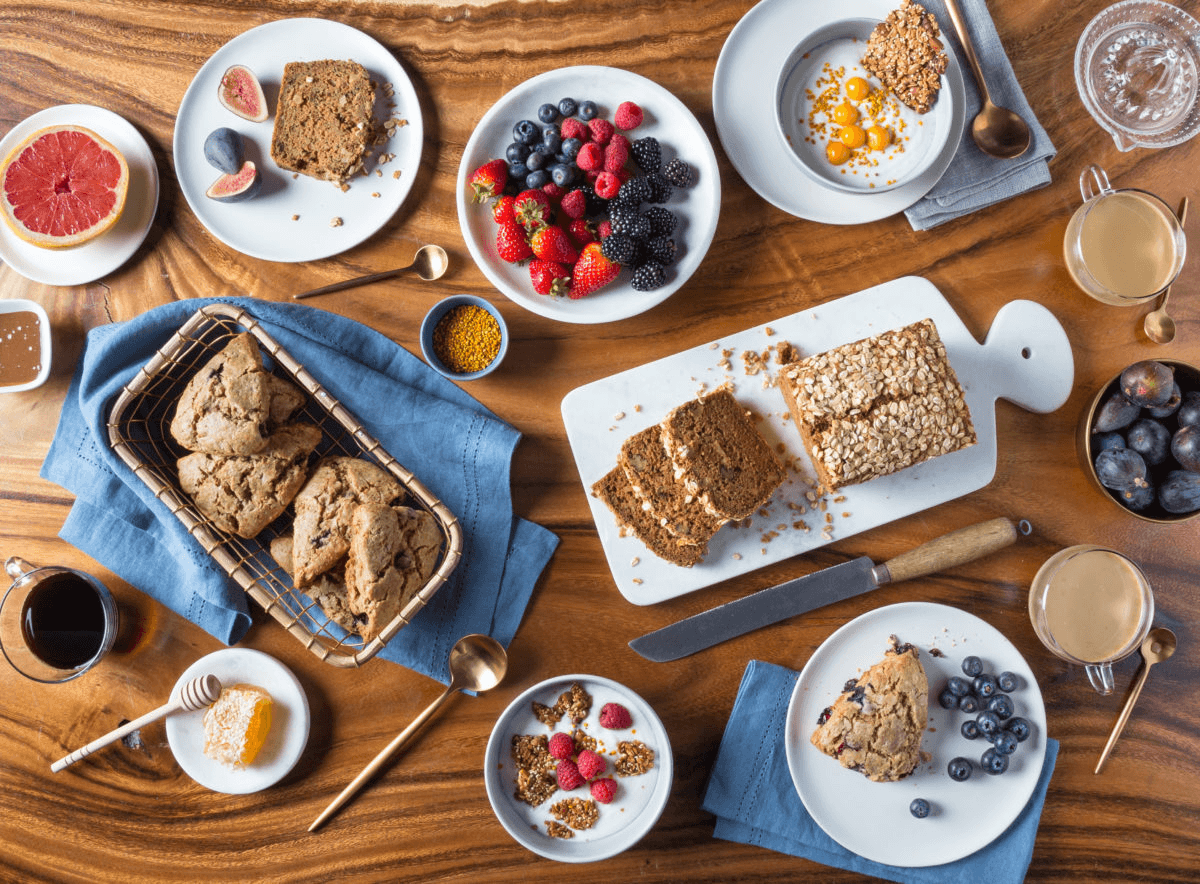 Aerial view of a breakfast spread on a wooden table, including gluten-free and refined sugar-free scones, loaf, berries, honeycomb, grapefruit halves, figs, and coffee.