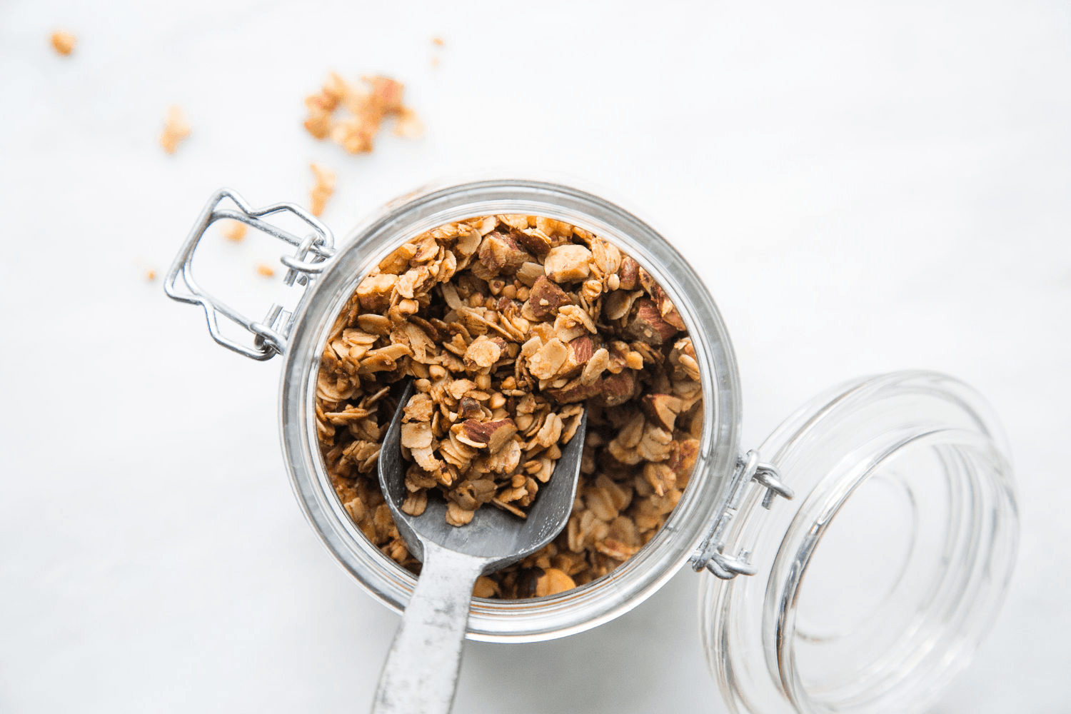 Aerial view of savoury granola in a glass mason jar with a silver spoon, and some granola scattered around on a white background.