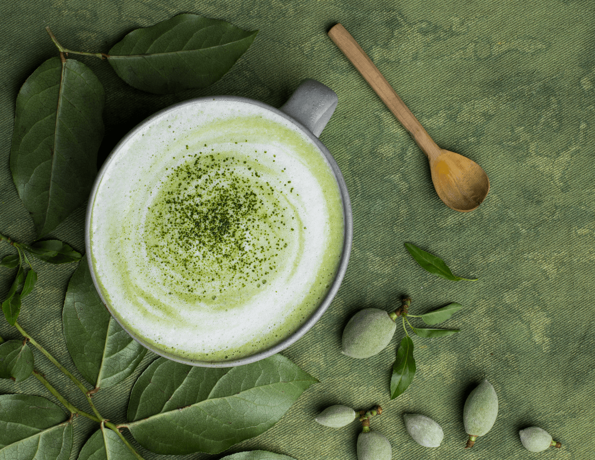 A frothy green matcha latte in a mug with sprinkled matcha on top, on a green backdrop next to green leaves and a wooden spoon.