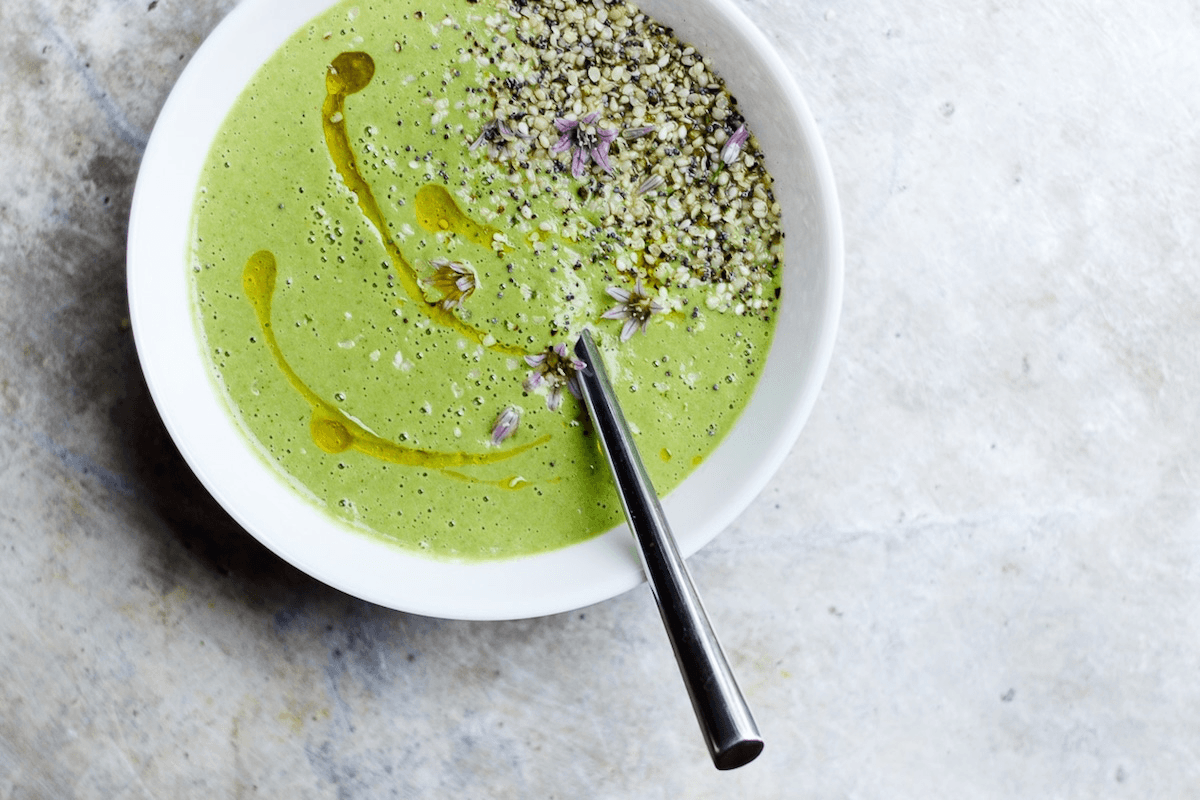 A spring green soup in a white bowl garnished with hemp seed sukkah and olive oil drizzle, with a silver spoon.