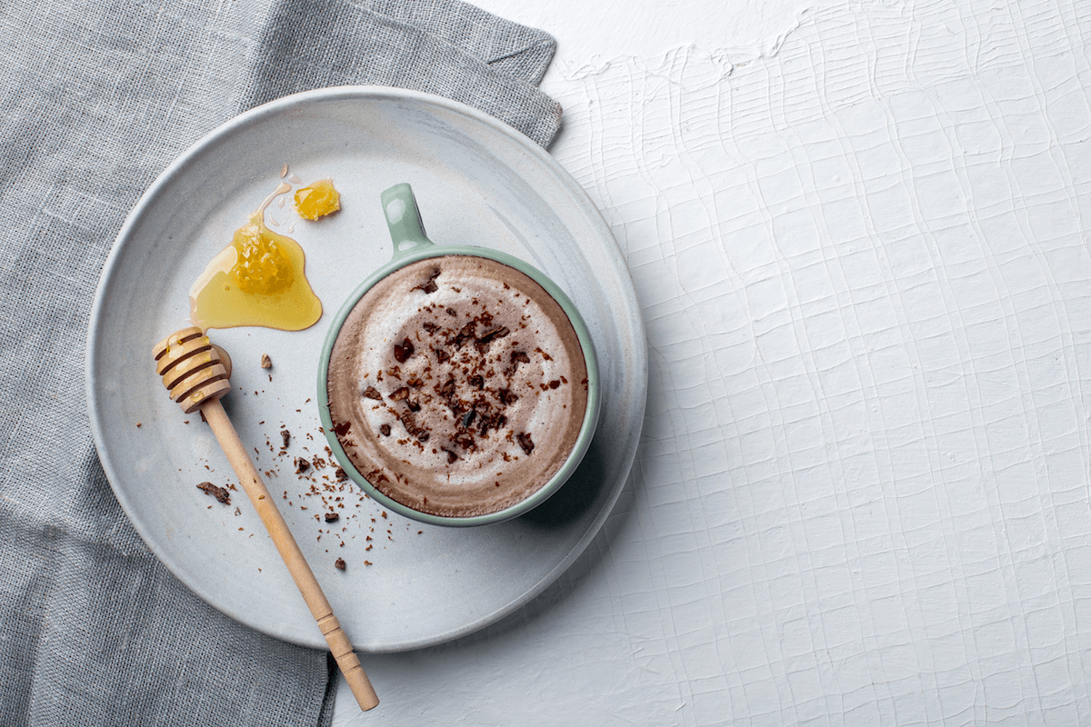 An aerial view of a frothy vegan hot chocolate in a blue mug on a white plate next to honey comb, sprinkled with chocolate shavings. 