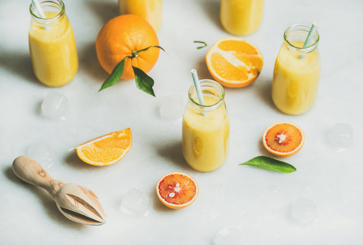 Small glass jugs of freshly squeeze orange juice with paper straws, surrounded by orange slices, orange leaves, and a lemon press.