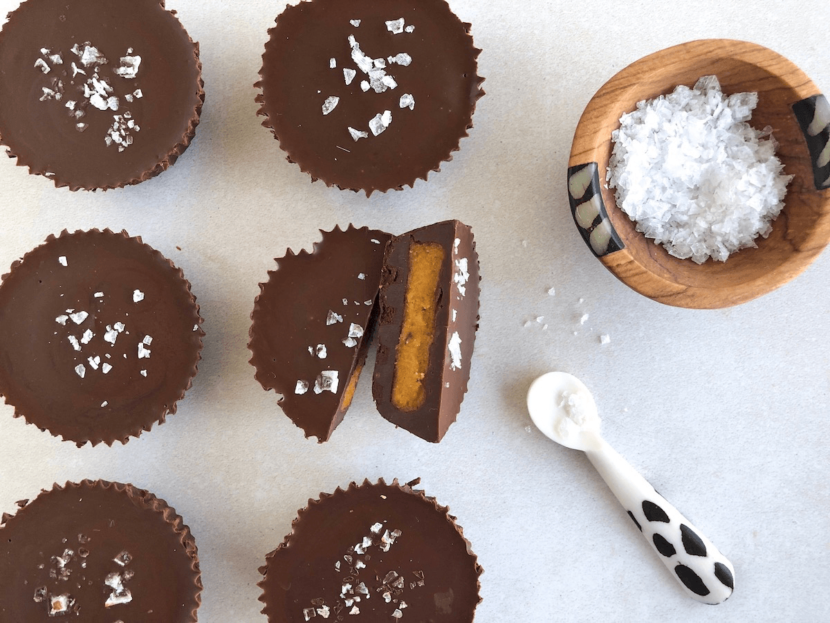 Aerial view of chocolate pumpkin peanut butter cups sprinkled with sea salt, with one cut in half to show the pumpkin center, next to a bowl of flaky sea salt.