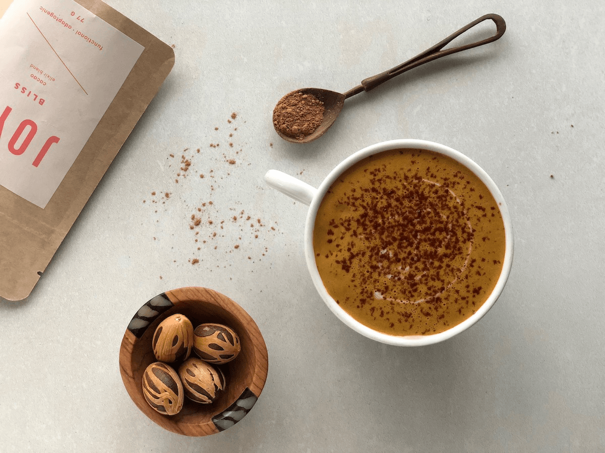 Aerial view of a homemade pumpkin spice latte in a white mug with cinnamon sprinkled on top, next to Joya's Bliss elixir packaging, a bowl of whole nutmegs, and a wooden spoon. 