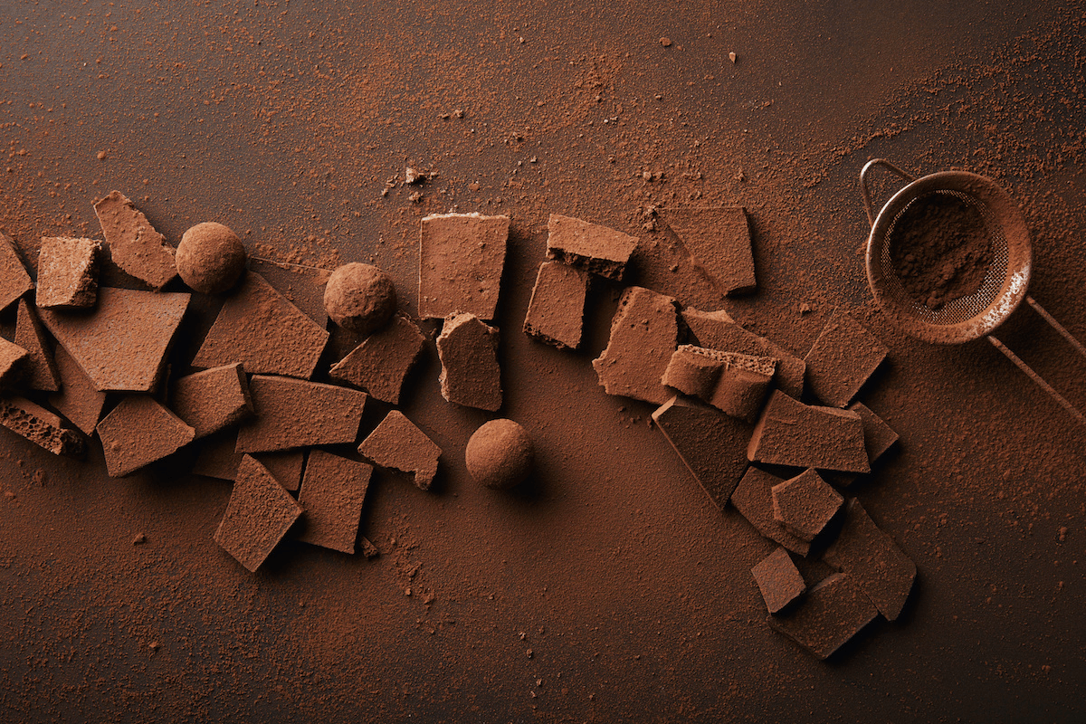 Aerial view of broken chocolate bars and chocolate truffles scattered across a dark backdrop, covered in raw cacao powder, next to a sieve full of raw cacao powder.