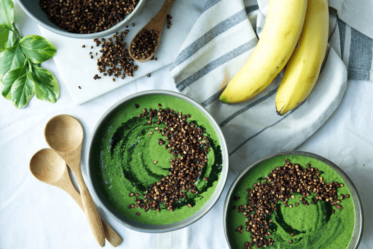 Two green smoothie bowls with a swirl of spirulina powder, topped with buckwheat crisps, next to two wooden spoons, bananas, and a large bowl of buckwheat crisps.