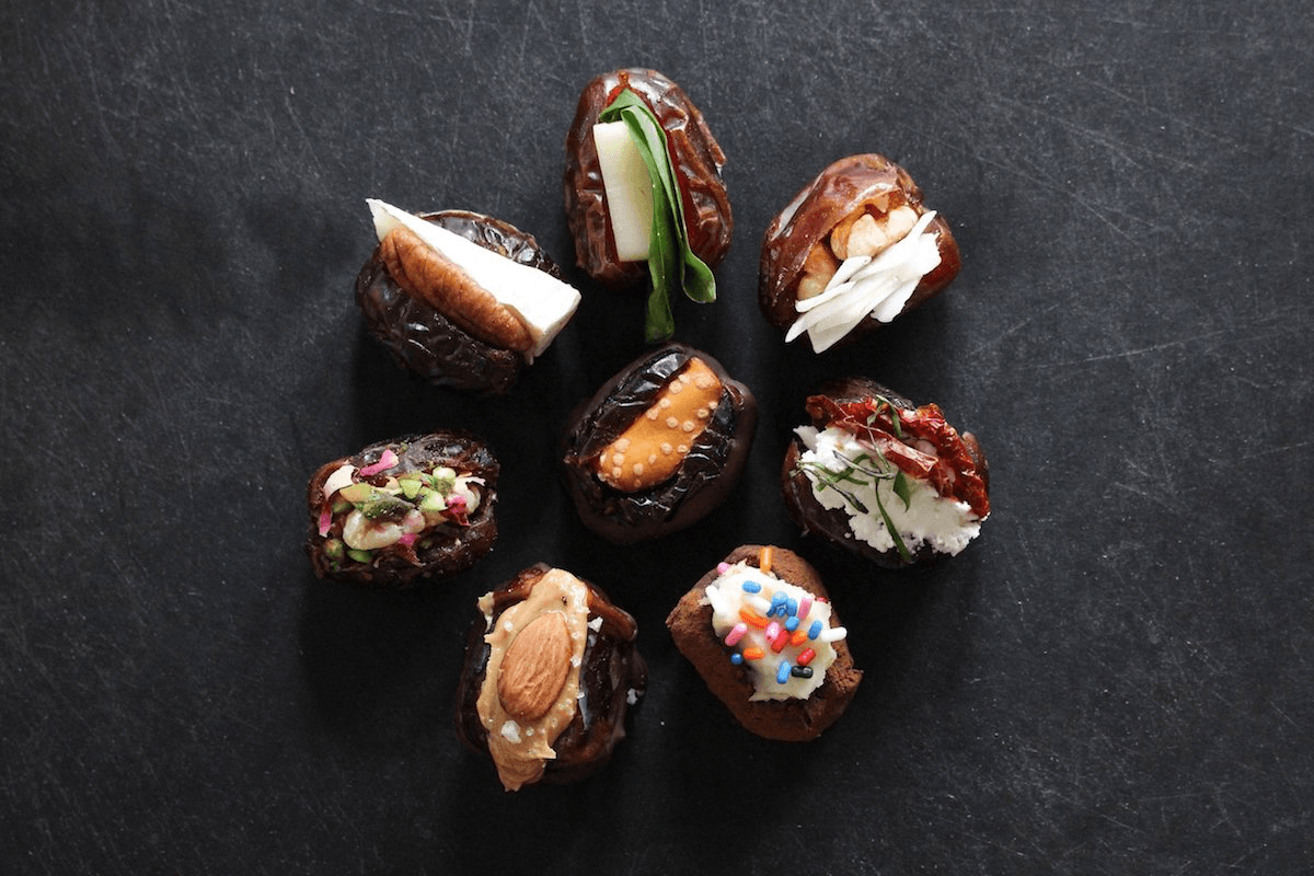 Dates in a circular formation on a black backdrop, stuffed with various ingredients like nut butter, cheese, pickles, roasted tomatoes, and herbs. 