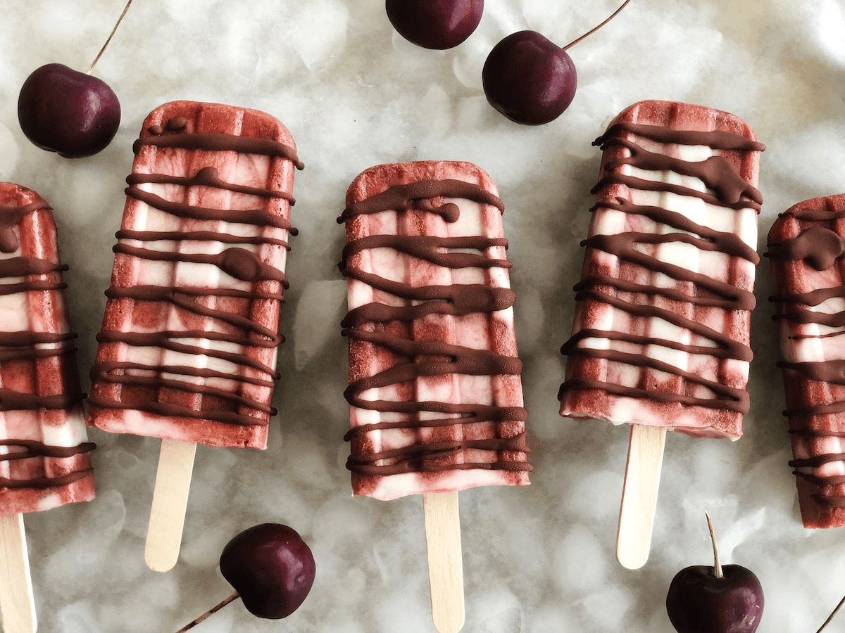 Five vegan cherry carcia creamsicles lie on a bed of ice drizzled with paleo chocolate and surrounded by black cherries. 