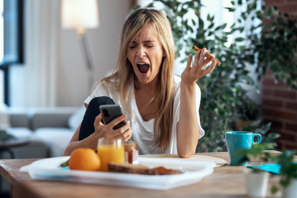 Blond woman yawning while eating breakfast and looking at her cellphone