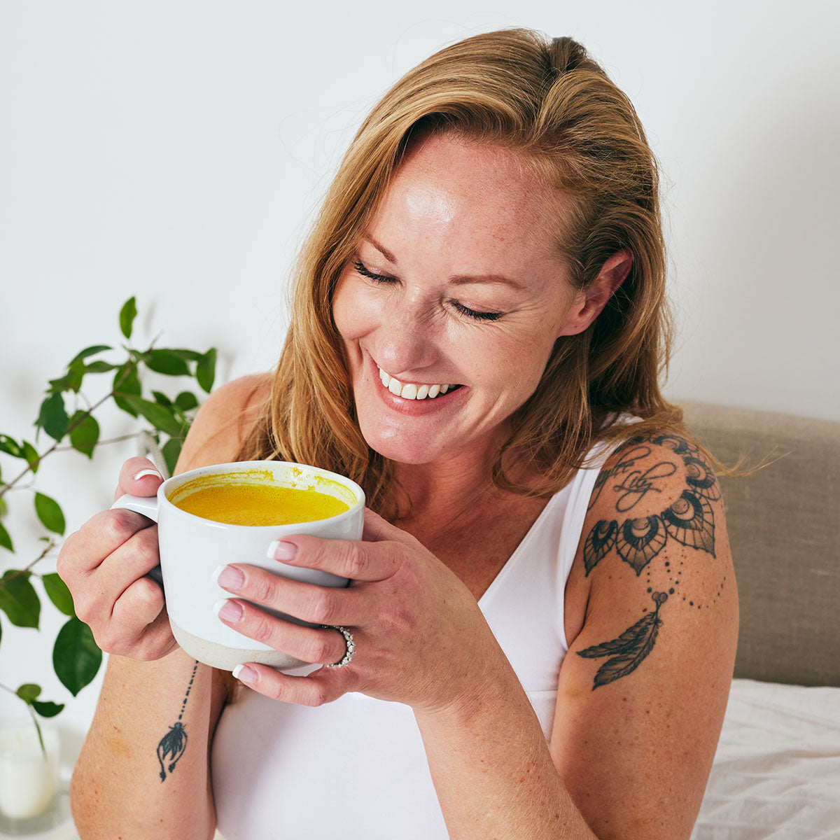 Woman sitting in bed smiling and holding a hot Restore turmeric latte in a white mug