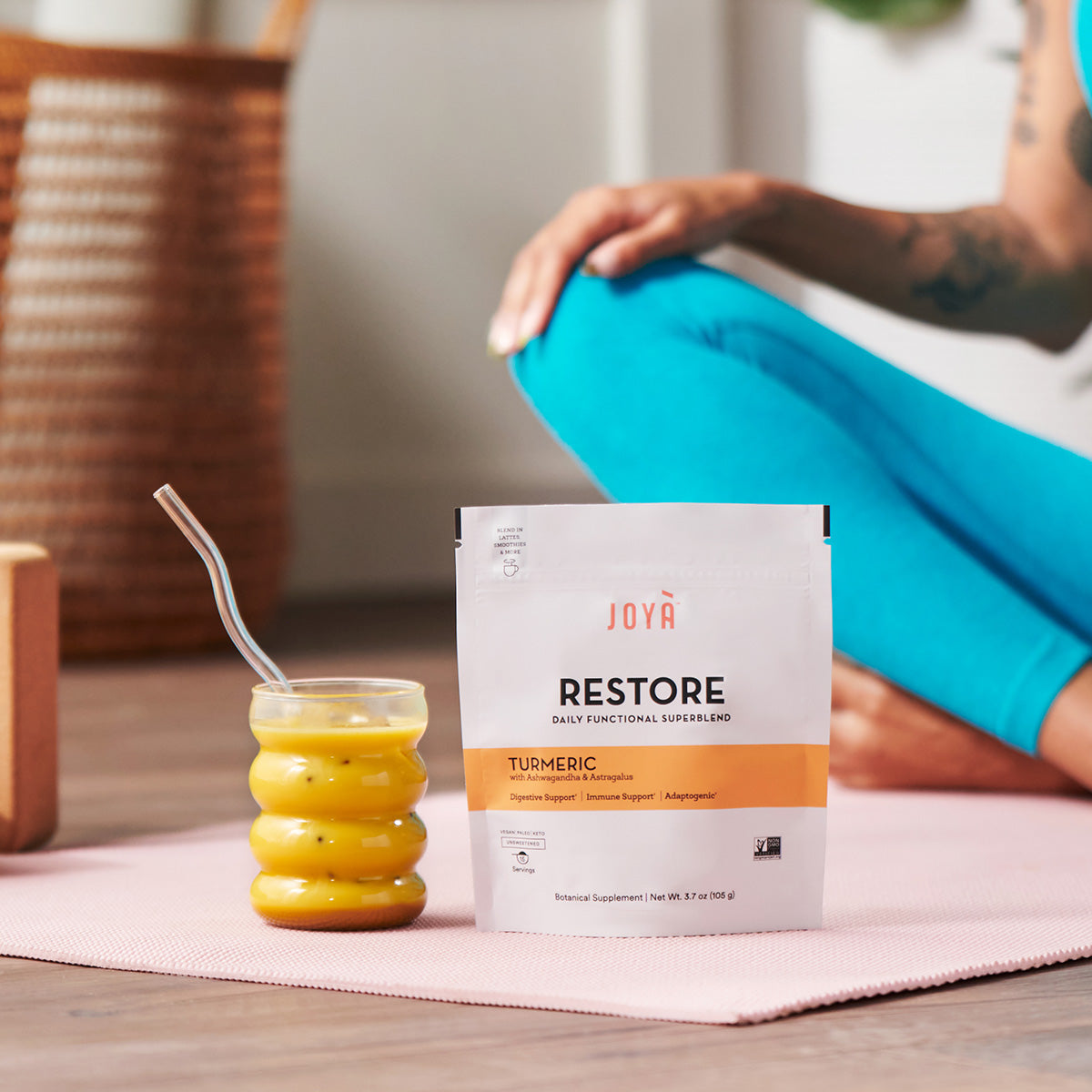 Restore Superblend beside an iced Restore turmeric latte on a yoga mat while a young woman meditates in the background