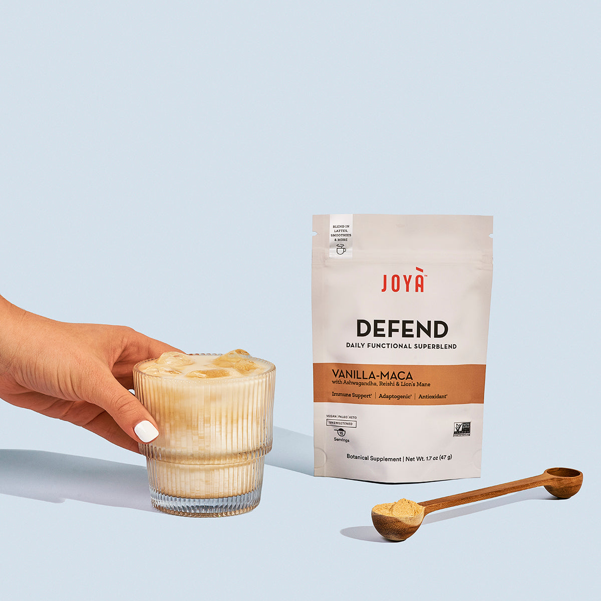 Defend Superblend 15-serving pouch sitting next to wooden spoon and a young woman's hand reaching for a glass of iced Defend vanilla-maca latte