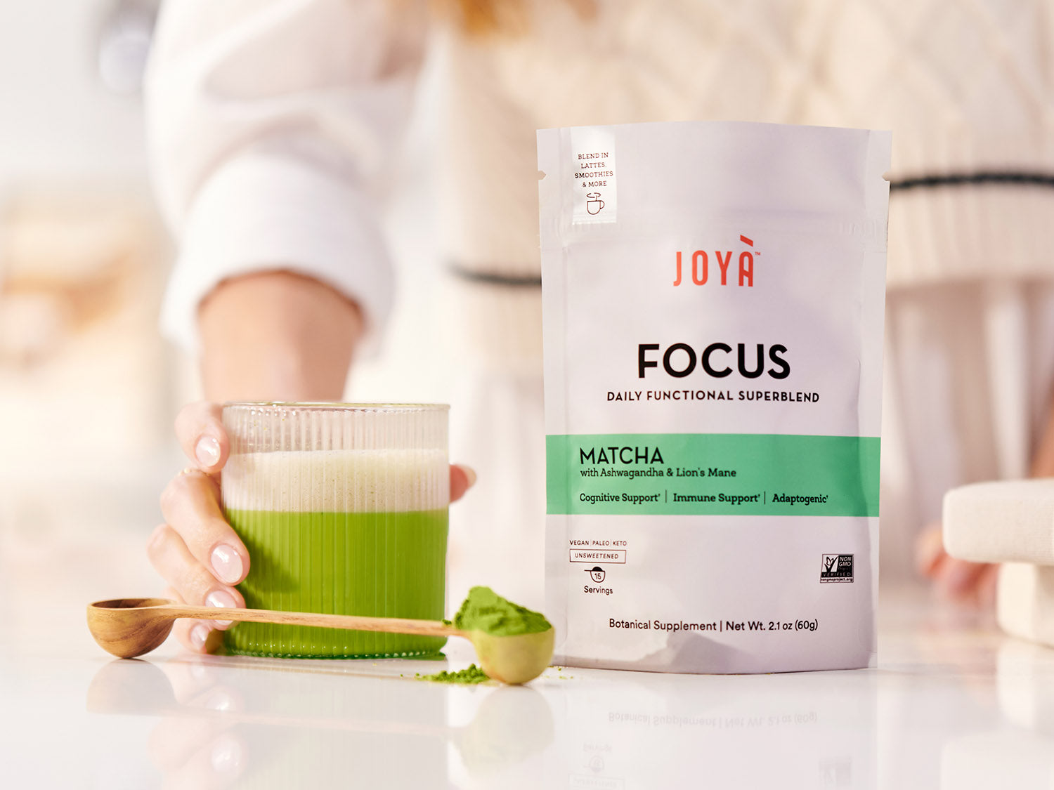 Focus Superblend 15-serving pouch sitting on a kitchen countertop beside a wooden spoon and a woman holding a hot matcha latte in a clear cup